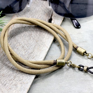 May include: A khaki-colored paracord eyeglass strap with brass hardware and black rubber ends.