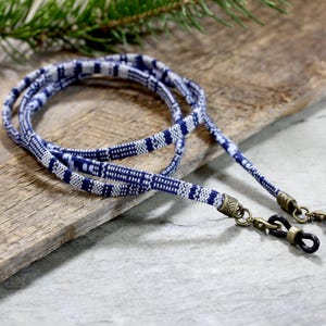 May include: A pair of blue and white patterned eyeglass holders with gold-tone metal clasps. The holders are made of woven fabric and are lying on a wooden surface.