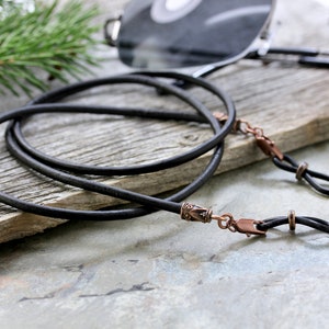 May include: Black leather eyeglass chain with copper-toned metal clasps and decorative beads. The chain is lying on a wooden surface.