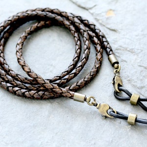 May include: Braided brown leather eyeglass chain with antique brass-colored connectors. The chain is looped and lies on a gray stone surface. The ends have black rubber loops to attach to glasses.