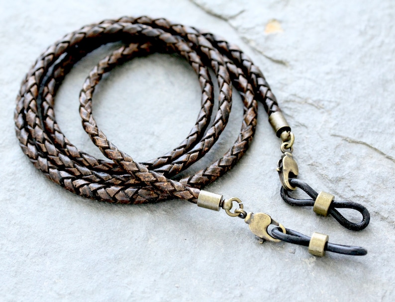 May include: Braided brown leather eyeglass chain with antique brass-colored connectors. The chain is looped and lies on a gray stone surface. The ends have black rubber loops to attach to glasses.