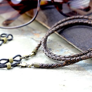 May include: Braided brown leather eyeglass chain with brass-colored clasps and connectors. The chain is lying on a textured surface next to a pair of sunglasses and two additional eyeglass connectors.