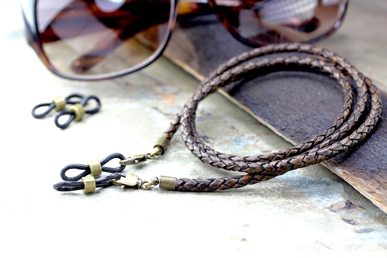 May include: Braided brown leather eyeglass chain with brass-colored clasps and connectors. The chain is lying on a textured surface next to a pair of sunglasses and two additional eyeglass connectors.