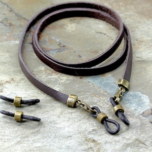 May include: A brown leather eyeglass chain with gold-tone metal clasps and black rubber tips. The chain is coiled and laid out on a textured surface.