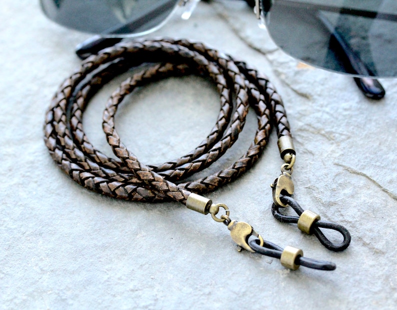 May include: Braided brown leather eyeglass chain with brass-colored clasps. The chain is looped and lies on a light gray surface, with a pair of sunglasses in the background. The chain is designed to hold eyeglasses.