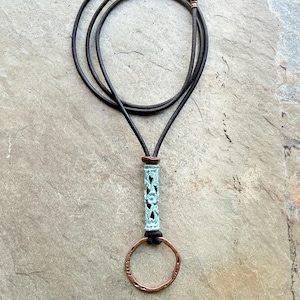 May include: A necklace featuring a dark brown leather cord with a copper-toned clasp. The pendant consists of a cylindrical, teal-colored bead with an intricate floral cutout pattern, suspended above a circular, hammered copper-toned metal ring.