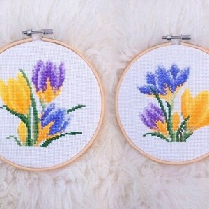 Puede incluir: Dos aros de bordado con diseños de crocus morados y amarillos en punto de cruz. Los aros son de madera y tienen un fondo blanco.