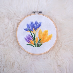 May include: A close-up of a cross-stitch embroidery hoop featuring a floral design. The design depicts purple, blue, and yellow crocuses.