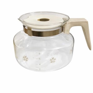 May include: A clear glass coffee carafe with a beige handle and lid. The carafe has a gold-colored band around the middle and floral designs. Measurements are marked on the side, up to 8 cups.