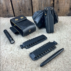 Op de afbeelding: Een verzameling zwarte gereedschappen en accessoires, waaronder een multitool, een leren etui, een zaklamp, bithouders en een pen. De items zijn gerangschikt op een getextureerd, gebroken witte ondergrond met een houten achtergrond.