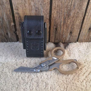 Op de afbeelding: Een zwart leren etui met een gestikt ontwerp en twee drukknopen. Het etui bevat een zwarte pen en een klein multitool met een beige handvat. Het multitool is gemarkeerd met "Leatherman" en "Raptor".