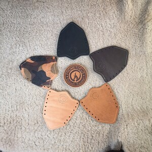 May include: An overhead shot displays several leather arm guards in various colors, including black, brown, and camouflage. A circular patch with the text "PROMETHEUS Bushcraft Equipment" is in the center. Each guard has a series of holes along the edges.