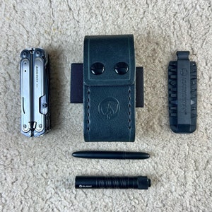 May include: A collection of black and silver tools and accessories, including a multi-tool, a leather sheath, a bit holder, a pen, and a flashlight. The multi-tool is silver with black accents. The leather sheath is dark green with a button closure.