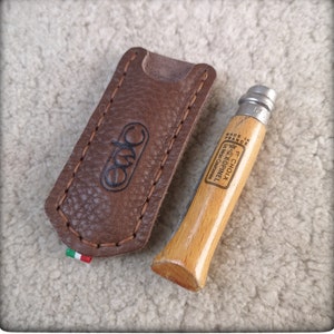 Puede incluir: Una funda de cuchillo de cuero marrón con un borde cosido y la palabra "Cuda" en relieve en la parte delantera. Un cuchillo de bolsillo con mango de madera y hoja de plata y el texto "Le  Couteau  Opinel  La  Main  Chauvignonne  Made in France" está al lado de la funda.
