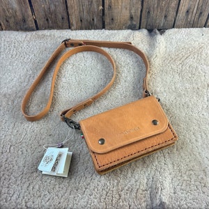 Könnte beinhalten: Eine braune Leder-Umhängetasche mit einem langen Riemen. Die Tasche hat eine Klappe mit einem Druckknopfverschluss und den Text "PROMETHEUS" auf der Vorderseite geprägt.