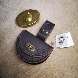 Könnte beinhalten: Eine braune Ledertasche mit Klappe und einem Messingverschluss. Die Tasche ist an einem braunen Lederriemen mit einer Messingschnalle befestigt. Die Tasche hat ein genähtes Design und ein Logo auf der Vorderseite. Die Tasche ist mit "Prometheus Equipment" beschriftet.