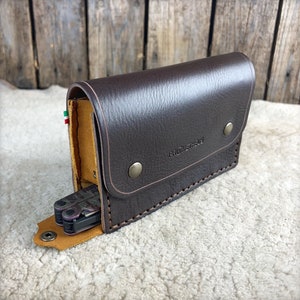 May include: A brown leather case with a stitched edge and a snap closure. The case is open to reveal a multi-tool with a yellow and green flag.