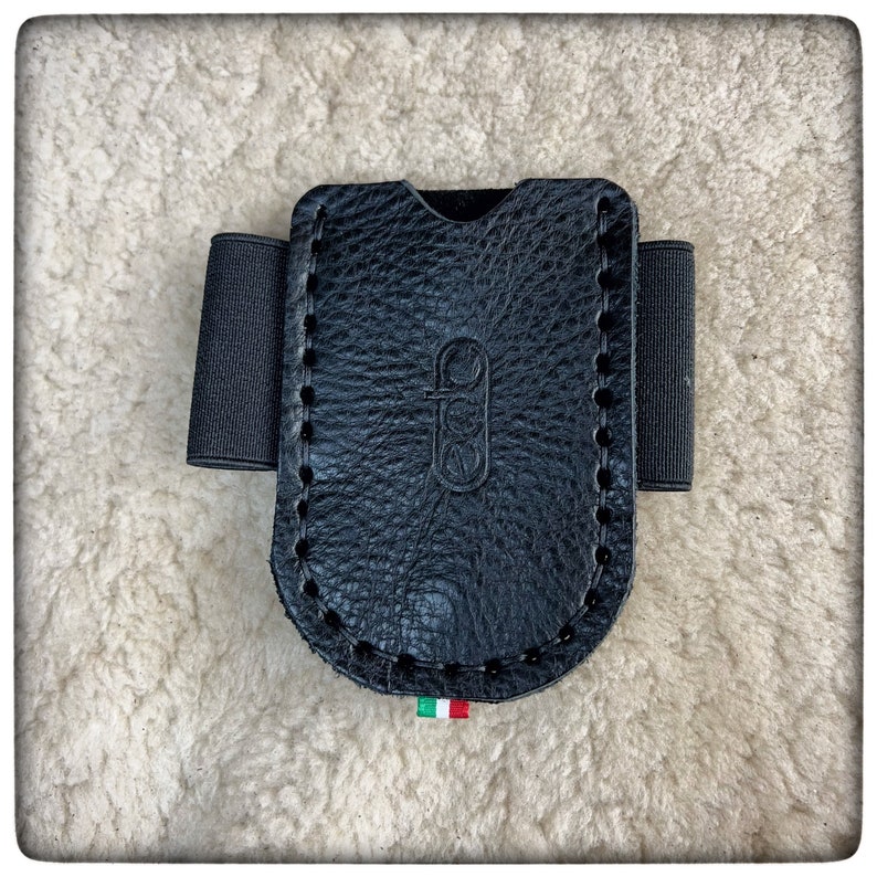 May include: Black leather card holder with a stitched edge and elastic wrist strap. The holder has a logo embossed on the front.