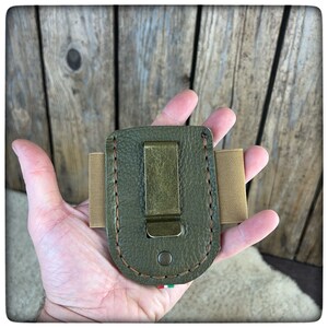May include: A green leather pocket clip with a brown stitched border and a metal clip. The clip is held in a hand.