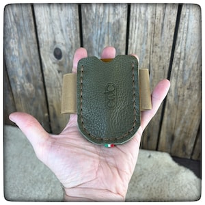 May include: A green leather card holder with a brown elastic band and a stitched design. The holder has a small Italian flag on the bottom and the letters "CCC" embossed on the front.