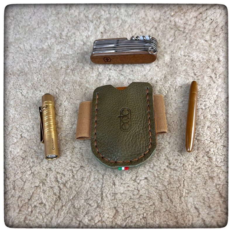 May include: A green leather EDC (Every Day Carry) wallet with a brown leather strap and stitching. The wallet has a small Italian flag on the bottom. A small gold flashlight and a multi-tool are also pictured.