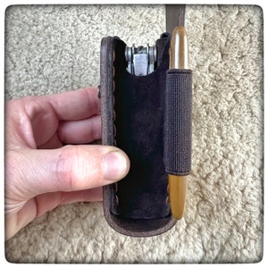 May include: A brown leather case with a zipper closure, designed to hold a multi-tool and a pen. The case has a suede lining and a stitched edge.