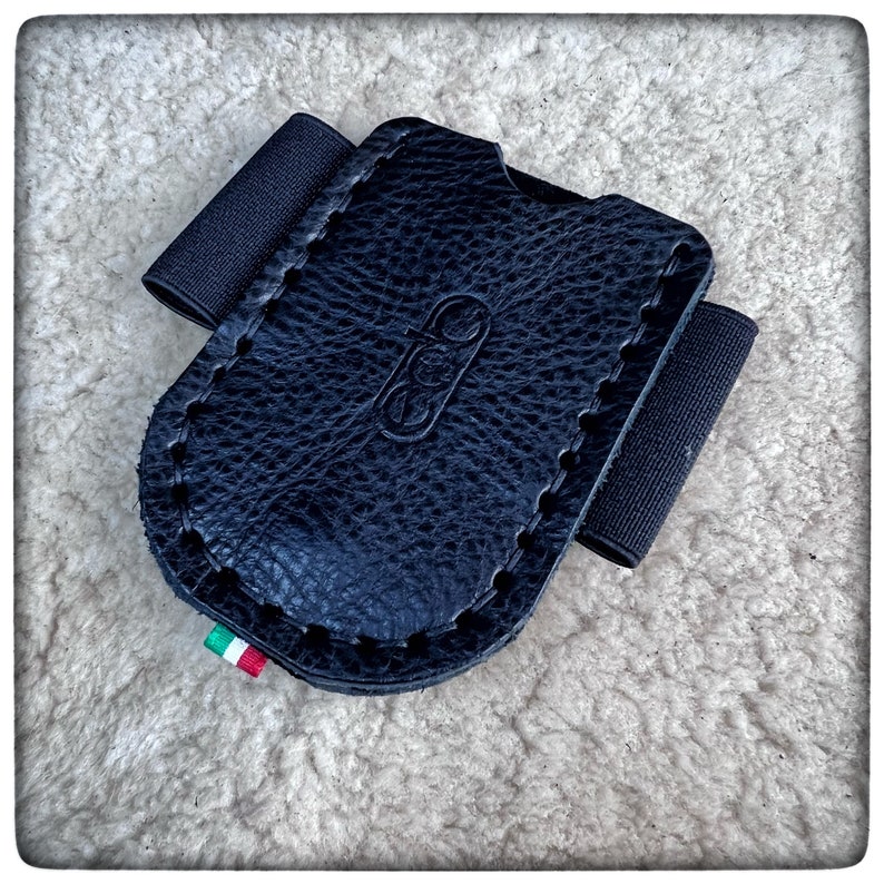 May include: Black leather card holder with a black elastic band and a stitched edge. The holder has a small Italian flag sewn on the bottom.