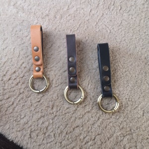 Può includere: Tre portachiavi in ​​pelle con anelli in metallo. I portachiavi sono marroni, neri e beige. Ogni portachiavi ha tre rivetti in ottone.