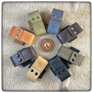 May include: Assortment of eight leather pouches in various colors, including brown, tan, green, and gray. Each pouch has a snap closure and a small logo embossed on the front. The pouches are arranged in a circle on a white fuzzy surface. The logo reads "Prometheus Bushcraft Equipment."