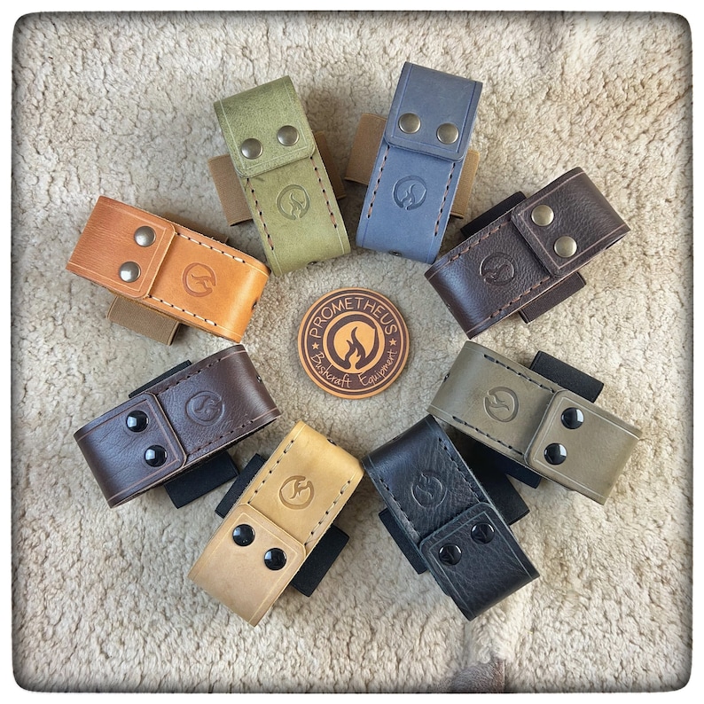 May include: Assortment of eight leather pouches in various colors, including brown, tan, green, and gray. Each pouch has a snap closure and a small logo embossed on the front. The pouches are arranged in a circle on a white fuzzy surface. The logo reads "Prometheus Bushcraft Equipment."