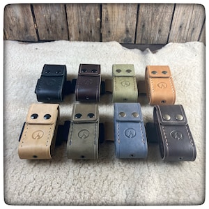 May include: Eight leather pouches in different colors, including black, brown, tan, green, blue, and gray. Each pouch has a stitched design and two metal snaps. The pouches are arranged in a row on a light brown surface.