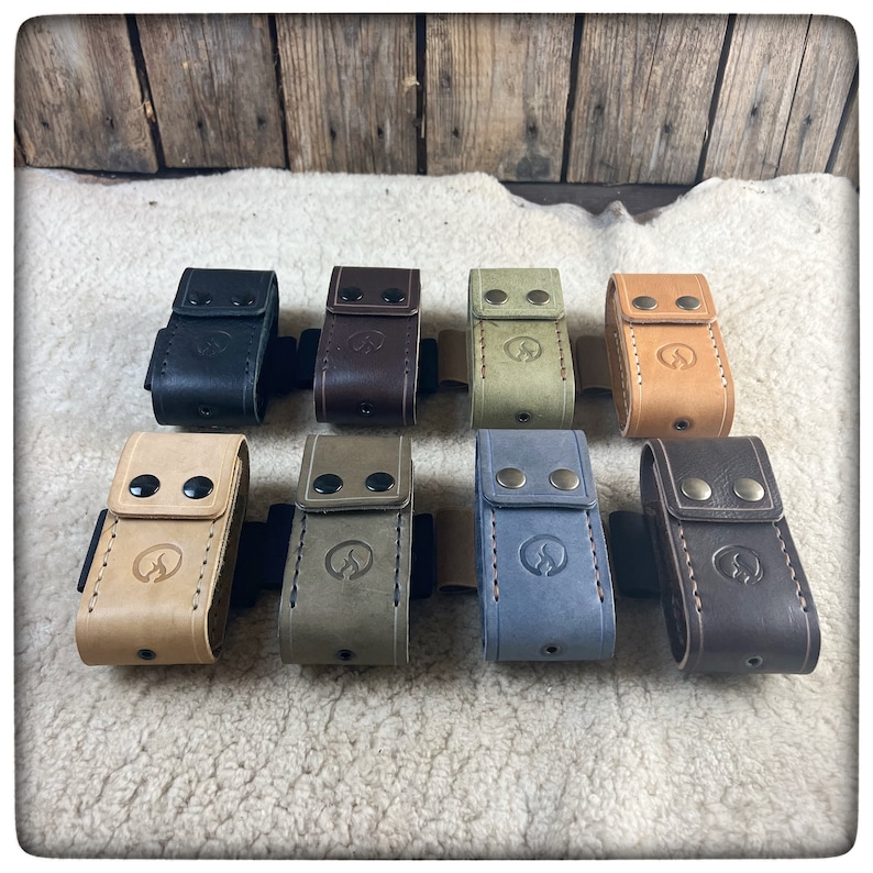 May include: Eight leather pouches in different colors, including black, brown, tan, green, blue, and gray. Each pouch has a stitched design and two metal snaps. The pouches are arranged in a row on a light brown surface.