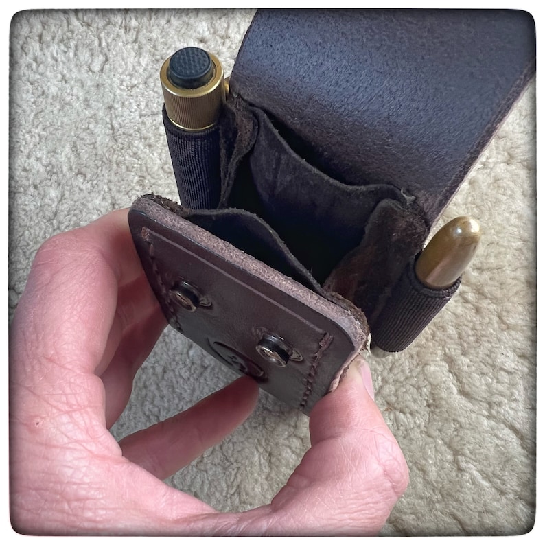 May include: A brown leather pouch with two compartments, one holding a small brass and black pen and the other holding a brass bullet. The pouch is open and the interior is visible.