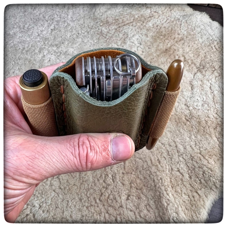 May include: A green leather pouch with a brown strap and a multi-tool inside. The pouch is held in a hand.
