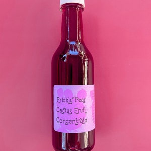 May include: A glass bottle of prickly pear cactus fruit concentrate. The bottle is filled with a deep purple liquid and has a white cap. The label is pink and says "Prickly Pear Cactus Fruit Concentrate".