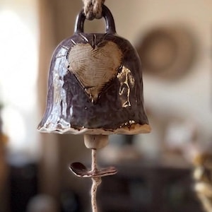 May include: A purple glazed ceramic bell hanging from a thick jute rope. The bell features a textured heart design on the front. A thin string hangs from the centre, adorned with a small ceramic bead and a dark purple heart-shaped charm.