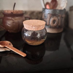 May include: A small, round, dark brown jar with a cork lid. A small wooden spoon rests on a small, round, light brown stone. A brown mug with a cork lid is in the background.