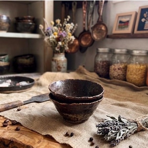 May include: Two stacked, dark brown, hand-thrown ceramic bowls with a textured, rustic finish. The bowls feature an uneven, organic rim and a deep, mottled glaze. They are positioned on a piece of burlap fabric alongside a metal spatula.