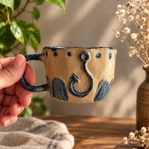 Puede incluir: Una taza de cerámica hecha a mano con base color canela y detalles en azul oscuro. La taza presenta un diseño de anzuelo en relieve en el centro, rodeado de puntos azul oscuro a lo largo del borde y patrones verticales de hojas en la base.