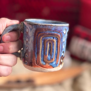 May include: A ceramic mug with a textured surface featuring a raised rectangular spiral design. The glaze is a blend of deep blue, metallic bronze, and earthy brown tones, with a series of small circular indentations along the side of the handle.