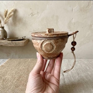 Puede incluir: Un cuenco de cerámica beige, pequeño y redondo, con tapa a juego y un acabado texturizado y moteado. El cuenco presenta un diseño circular en relieve en el lateral y un asa con un cordel adornado con dos pequeñas cuentas de madera marrón oscuro.