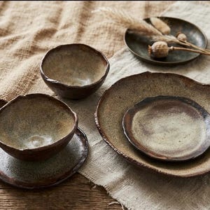 Puede incluir: Un conjunto de vajilla de cerámica hecha a mano que incluye dos cuencos y tres platos con bordes irregulares y orgánicos. Las piezas tienen un acabado texturizado y moteado en tonos tierra, marrón, beige y gris, con un aspecto rústico.