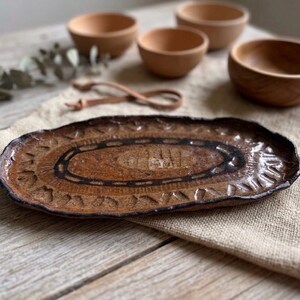 Puede incluir: Una bandeja ovalada de cerámica con una superficie marrón texturizada que presenta patrones grabados en marrón oscuro y un borde festoneado. La bandeja se acompaña de cuatro cuencos de madera pequeños, lisos y de color marrón claro.