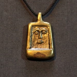May include: A gold pendant necklace with a stylized face design. The pendant is rectangular and has a textured surface.
