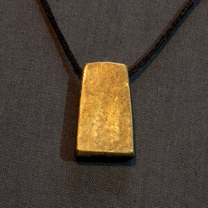 Puede incluir: Un colgante de color dorado en forma de rectángulo cuelga de un cordón negro.