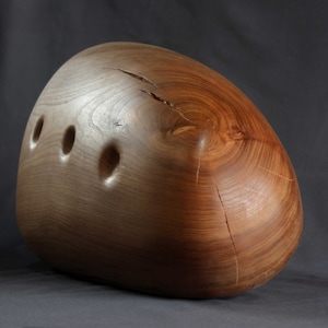 May include: A smooth, wooden sculpture with a rounded, organic shape. The sculpture has a natural wood grain and three holes carved into its surface.