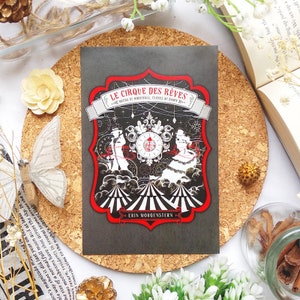 Könnte beinhalten: Ein Buchcover mit schwarzem Hintergrund und einem roten und weißen Zirkuszeltdesign. Der Titel des Buches lautet "Le Cirque des Rêves" und der Autor ist "Erin Morgenstern".