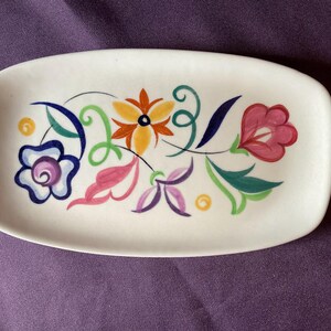 Puede incluir: Una bandeja ovalada de cerámica blanca con un diseño floral pintado a mano. El patrón incluye una flor central amarilla y naranja, una flor azul y morada, una flor rosa y varias hojas y espirales de colores verde, rosa y morado.
