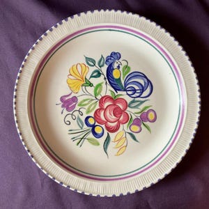 Peut inclure: Une assiette ronde en céramique avec un centre crème et un bord côtelé orné de points bleus. Le centre présente un motif floral peint à la main avec un coq bleu, une grande rose rouge, des fleurs jaunes et violettes, et des feuilles vertes.