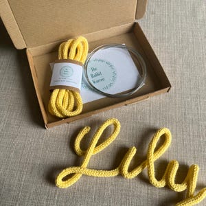 May include: A kit for making a knitted name sign. The kit includes yellow yarn, a metal wire form, and a label that reads "The Rabbit Warren". The name "Lily" is knitted in yellow yarn.